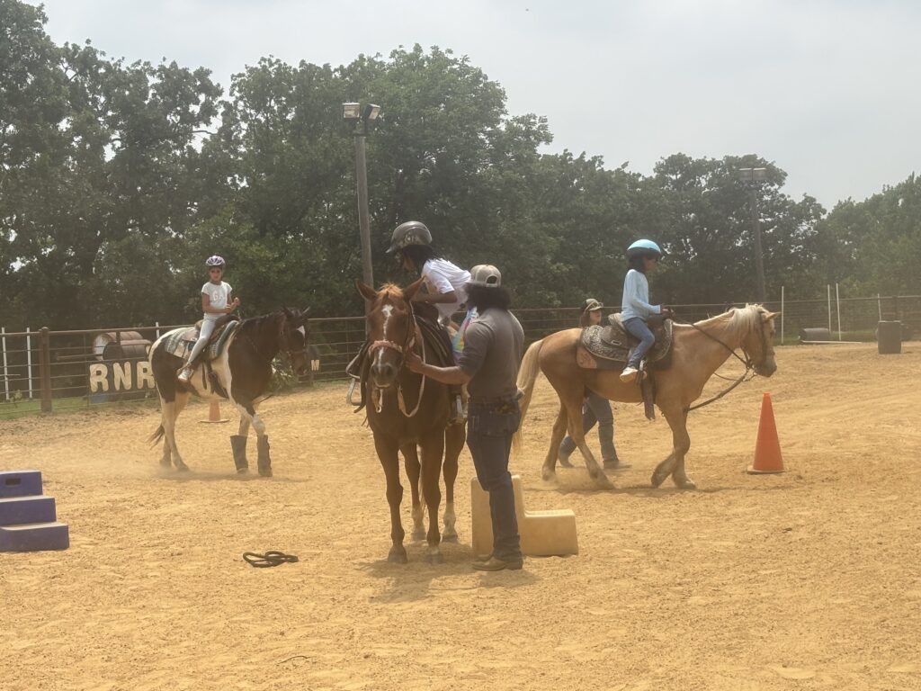 Children riding horses in a sandy outdoor arena with cones and trees.